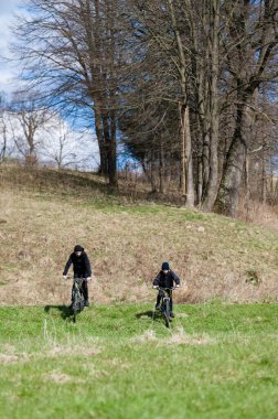 Bisikletli iki birey, hafif eğimli çimenli bir alanda karşılaşır. Arka planda yapraksız ağaçlar var. Bu da sonbahar ya da bahar olduğunu gösteriyor..