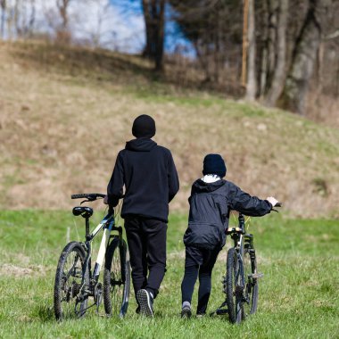 Arkadan görülen iki genç insan, çimenli bir tepe boyunca bisikletleriyle yürüyorlar. Kalın giyinmişler ve ayarı bahar ya da sonbahar günü olduğunu gösteriyor..
