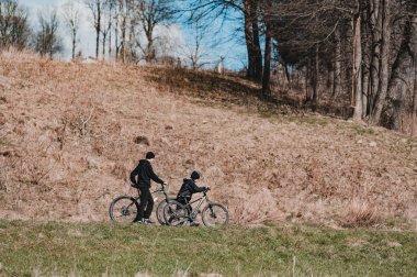 Bir yetişkin ve bir çocuğun güneşli bir günde çimenli bir yamaçta bisiklet sürdüğü sakin bir açık hava sahnesi. Mekan huzurlu ve huzurlu..