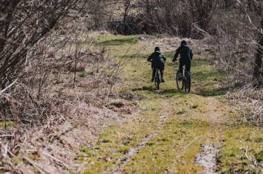 Bir çift bisikletçi, arkadan görülüyor, bir patikada yol alıyorlar. Yol yapraksız çalılar ve ağaçlarla çevrili, çimenli. Bu da sonbaharın sonları ya da ilkbaharın başları olduğunu gösteriyor..