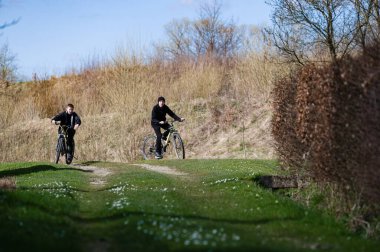 İki genç insan ilkbahar güneşinin tadını çıkararak bir kır yolunda bisiklet sürüyor. Yol bir tarladan geçiyor, çalıların ve ağaçların arasından, huzurlu ve manzaralı bir doğa deneyimi yaratıyor..