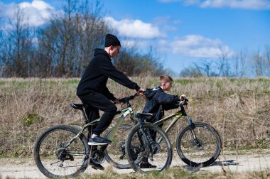 Güneşli bir günde bir erkek ve çocuk birlikte bisiklet sürerler. Yol toprak ve kuru otlar yolu çevreleyecek. Temiz havanın ve güneşin tadını çıkarıyorlar..
