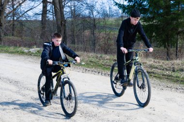İki genç çocuk bir kırsal şerit boyunca bisiklet sürüyor. Gün ışığı, açık hava macerasının tadını çıkarırken sahneyi aydınlatıyor. Yol toprak ve ağaçlar yol kenarında..