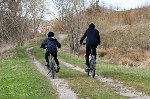 Bisikletli iki kişi çimenli bir arazide kirli bir patika boyunca bisiklet sürüyor. Ağaçlar ve çalılar yolu çiziyor. Barışçıl bir açık hava sahnesi.
