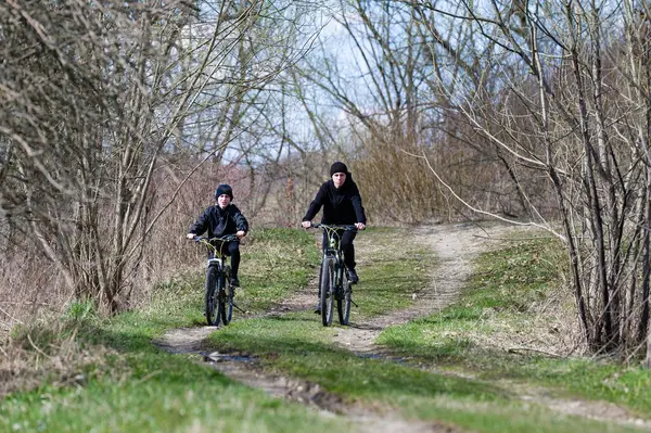 Bir çift birey, yapraksız ağaçlarla çevrili toprak bir patika boyunca bisiklet sürüyor. Sahne, ilkbaharın başlarında bir dinginlik ve açık hava aktivitesi hissi uyandırıyor..