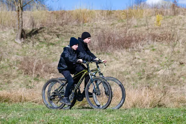 Parlak ve güneşli bir günde dağ bisikleti süren iki genç çocuk bir yol boyunca bisiklet sürüyor. Sıcak ceketler ve şapkalar giyip, dışarıda eğleniyorlar..