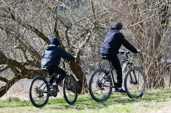 Bir patikada dağ bisikleti süren iki bisikletlinin manzaralı bir fotoğrafı. Çıplak ağaçlar sahneyi çevreleyip bahar gününe biraz kış katıyor..