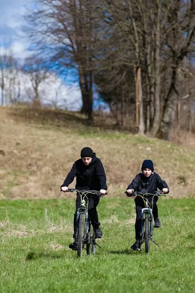 Açık mavi bir gökyüzünün altında canlı yeşil bir arazide bisiklet süren iki gencin resimli bir sahnesi. Arkaplanda bir sıra ağaç ve hafif eğimli bir tepe var..