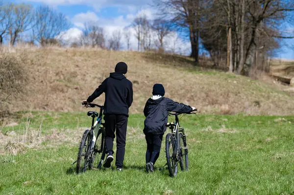 Bisikletleriyle uzaklaşan iki gencin dikiz aynasından. Parçalı bulutlu bir gökyüzünün altındaki yeşil çimen tarlasındalar. Arka planda ağaçlar ve tepeler var.