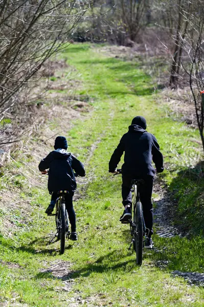 Bisikletli bir çift birey yeşil bir yolda yürür. İzler doğal bir manzara boyunca esiyor, sakin bir açık hava deneyimi sunuyor..