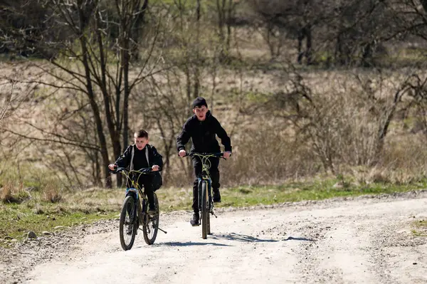 İki genç çocuk çakıllı bir yolda bisiklet sürüyor, doğal ortamda sakin bir gezinti yapıyorlar. Ağaçlar ve çalılar yol boyunca uzanır, açık hava aktiviteleri için manzaralı bir zemin sağlar..