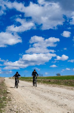 Çakıl yolu boyunca bisiklet süren iki bireyin manzaralı görüntüsü. Pofuduk beyaz bulutlarla kaplı parlak mavi gökyüzü, kırsal bölgelerde sakin bir gezinti için güzel bir zemin sağlar..