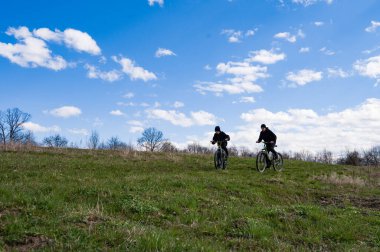 Güzel bir günde çimenli bir tepenin üzerinde bisiklet süren iki kişinin manzarası. Gökyüzü berrak ve parlak, kabarık beyaz bulutlarla. Sakin ve aktif bir açık hava sahnesi..