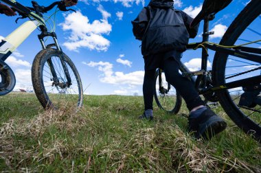 Çimenli bir tepeden dağ bisikletlerini iten iki kişinin alçak açılı görüntüsü. Gökyüzü kabarık beyaz bulutlarla parlak mavi. Sakin bir açık hava sahnesi..