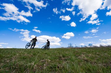 Geniş açı, dağ bisikletli iki kişiyi yeşil bir yamaçta yakalar. Pofuduk beyaz bulutlarla benekli açık mavi bir gökyüzünün arkaplanına karşı.