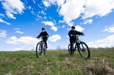 İki genç bisikletçi çimenli bir tepede dağ bisikleti sürüyor, açık mavi gökyüzü ve tüylü beyaz bulutlarla güzel bir günün tadını çıkarıyorlar. Açık hava macerası için mükemmel bir gün..