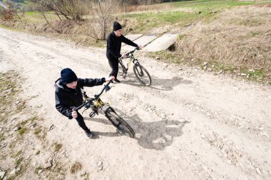 Genç figürler bisikletleriyle çakıllı bir yolda ilerliyorlar. Güneş ışığı manzarayı aydınlatıyor, huzurlu bir kırsal zemin sergiliyor..