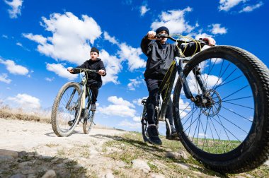 İki genç bisikletçi parlak mavi bir gökyüzünün altında toprak bir yol boyunca dağ bisikletlerine biniyorlar. Çocuklar mutlu ve enerjik görünüyorlar, açık hava maceralarının tadını çıkarıyorlar..