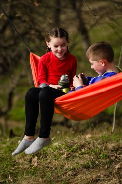 Kardeşler, sakin bir açık hava ortamının ortasında, parlak turuncu bir hamakta dinleniyorlar. Bir içkiyi paylaşırlar ve mutlu görünürler. Huzurlu ortamın tadını çıkarırlar..