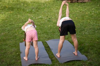 İki çocuk dışarıda minderlerde yoga pozu çalışıyor. Bir kız ve bir oğlan serin yeşil bir alanda esneme hareketleri yapıyorlar. Sağlıklı aktif yaşam biçimi.
