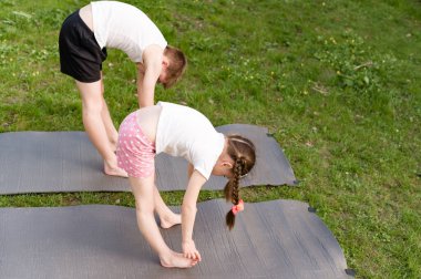 Çimenli bir parkta iki çocuk yoga minderleri üzerinde viraj yapıyor. Odaklanmışlar ve açık hava egzersizlerinin tadını çıkarıyorlar..