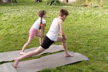 İki çocuk, bir erkek ve bir kız, çimenli bir bahçede yoga minderleri üzerinde esneme egzersizleri yapıyorlar. Odaklanmışlar ve antrenmanlarından zevk alıyor gibi görünüyorlar..
