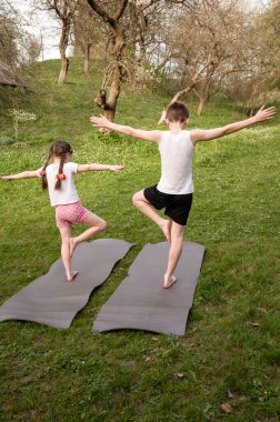 Çimenli bir alanda iki çocuk yoga pozu veriyor. Dengede duruyorlar, kollarını açıyorlar. Ortalık sakin ve huzurlu..