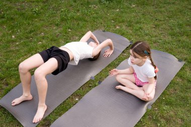 İki çocuk, bir erkek ve bir kız, gri paspaslarda yoga pozu veriyorlar. Çimenli bir bölgede güneşli bir günün tadını çıkarıyorlar..
