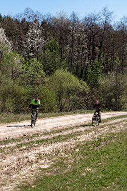 Toprak bir yolda bisiklet süren iki insanın manzarası. Ağaçlar ve yeşil yapraklar etraflarını sarmış, barışçıl ve açık hava ortamı yaratıyorlar..