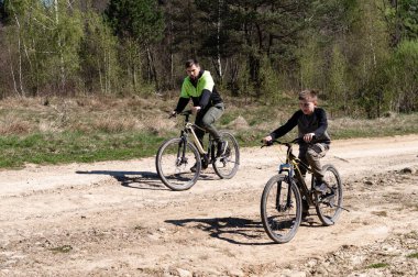 Bir adam ve bir çocuk kırsal bir yolda bisiklet sürüyorlar. Güneş ışığı, ormanlık bir alanda gezinirken manzarayı aydınlatır. Açık havanın tadını çıkarırlar..