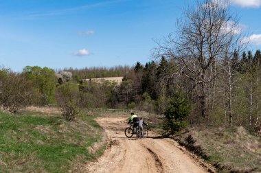 Bir çift bisikletçi toprak bir yolda pedal çeviriyor, doğal bir manzaranın dingin güzelliğine dalmış. Ağaçlar ve tarlalar yolu çerçeveleyerek, resim gibi bir ortam oluşturur.