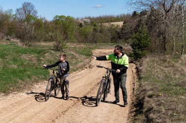 Bir adam ve bir çocuk kumlu bir yolda bisikletlerini yürütür. Parlak ve güneşli bir günde ağaçlar ve tarlalar sakin bir zemin oluşturur. Baba-oğul kaynaşma anı.