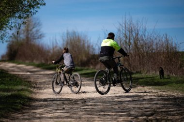 İki bisikletçinin, bir yetişkinin ve bir çocuğun güneşli bir günde keyifli bir yolculuğun keyfini çıkardıkları iç açıcı bir sahne. Çalı ve ağaçlarla kaplı toprak bir yoldan geçerler. Açık hava manzarası yaratırlar..