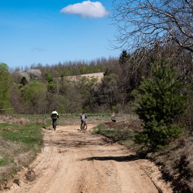 İki bisikletçinin bereketli bir arazide kumlu bir yolda sürüşünün sakin bir sahnesi. Güneş parlıyor, ağaçlar ve tarlalar onları kuşatıyor. Açık hava aktivitesi için mükemmel bir gün..