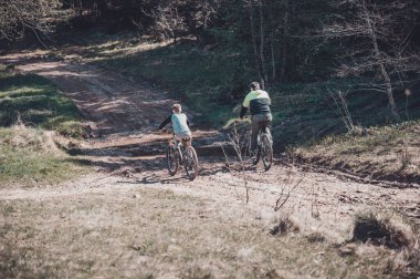 İki bisikletçi, muhtemelen bir baba ve oğul, kırsal bir yolda dağ bisikleti sürüyorlar. İzler ormanlık alanda esiyor, sakin bir açık hava deneyimi sunuyor..