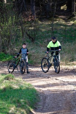 Bir adam ve çocuk dağ bisikletleriyle güneşli bir ormanda toprak bir patikada dikiliyorlar. Yolculuklarına ara vermiş gibi görünüyorlar..