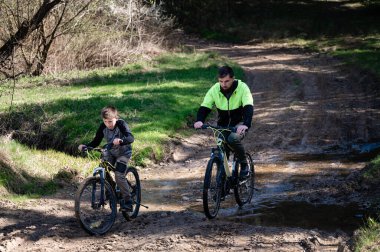 Bisikletli bir adam ve bir çocuk ağaçlık alanda çamurlu bir yolda ilerliyorlar. Küçük, sığ bir dereyi geçiyorlar. Sahne huzurlu ve macera hissi uyandırıyor..