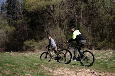 İki genç çocuk, yemyeşil ağaçlarla çevrili toprak bir patika boyunca bisiklet sürüyor. Güneş ışığı, orman tepe örtüsünün içinden süzülerek parlak ve maceracı bir atmosfer yaratır..
