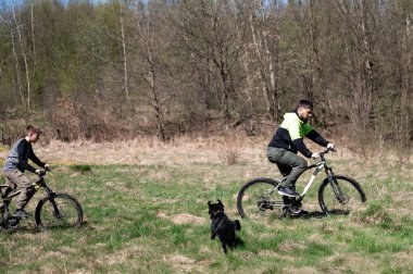İki bisikletçi, bir adam ve bir çocuk, çimenli bir arazide bisiklet sürerek güneşli bir günün tadını çıkarıyorlar. Siyah köpekleri onların yanında mutlu bir şekilde koşar, açık hava macerasının tadını çıkarırlar..