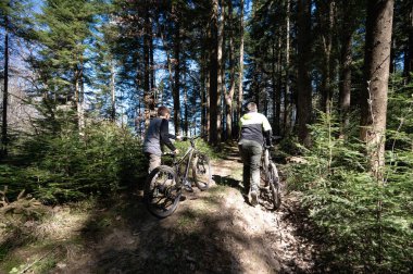 İki kişi sık bir orman boyunca güneş lekeli bir patika boyunca dağ bisikletlerini yürür. Uzun ağaçlar etraflarını sarar ve sakin bir atmosfer yaratır..