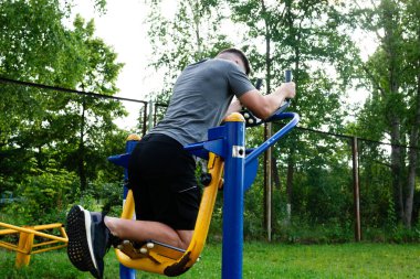 Bir insan arka tarafta spor yaparken açık hava fitness makinesi kullanır. Sahne yeşil çimenli ve ağaçlı bir park. Ekipman basit ve sağlam..