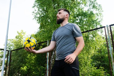 Gri gömlekli sakallı bir adam paslı bir çitin yanında sarı ve mavi bir futbol topuyla hokkabazlık yapıyor. Arka plan yemyeşil ağaçlar..