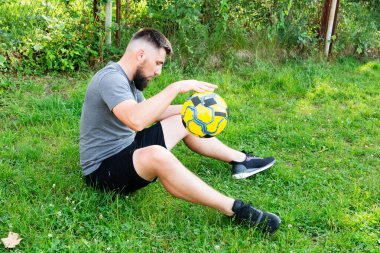 Sakallı bir adam çimenli bir alanda oturur, sarı ve siyah bir futbol topunu dikkatle inceler. Spor kıyafetleri giymiş, spor aktivitelerine ara verdiğini söylüyor..