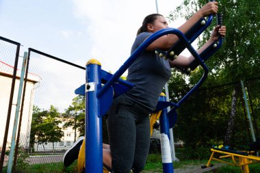 Bir kadın açık hava spor aletleri üzerinde güçlü bir eğitim egzersizi yapıyor. Ortam bir park ya da açık hava eğlence alanı. Atletik kıyafetler giyiyor..