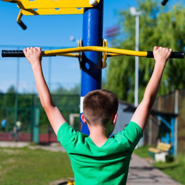 Arkadan genç bir çocuk açık hava barfiks çubuğu kullanırken görülüyor. Güneşli hava ve yeşil çevre, zindelik ve refah hissini arttırır..