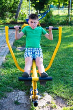 Genç bir çocuk idmanına odaklanmış sarı bir egzersiz makinesi kullanıyor. Güneşli bir parkta yeşil gömlek ve kareli şort giyiyor..