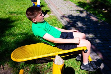 Genç bir çocuk açık hava egzersiz ekipmanlarında mekik çekiyor. Parlak sarı bank yeşil çimlerle ve çocuğun yeşil gömleğiyle çelişiyor. Parkta güneşli bir gün.
