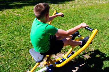 Genç bir çocuk açık hava egzersiz ekipmanları üzerinde güneşli bir çalışma yapıyor. Sarı bir makineye oturmuş, aktif olarak spor yapmak için kullanıyor..