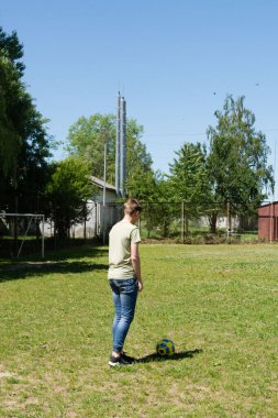Güneşli bir günde genç bir insan futbol topunu tekmelemeye hazır duruyor. Dekor, muhtemelen bir çitin ve bazı binaların yakınında bir açık araziye benziyor..