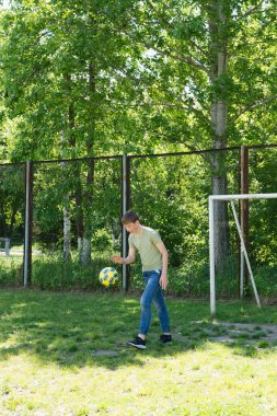 Genç bir çocuk çimenli bir arazide futbol topuyla hokkabazlık yapıyor. Arka planda bir futbol golü görünür. Ağaçların arasından güneş ışığı akıyor..